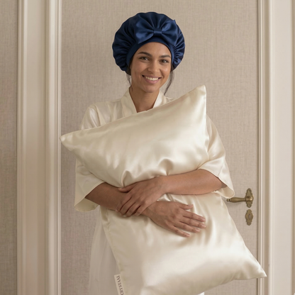 femme portant un bonnet de nuit en soie 22 momme bleu marine et tenant une taie d’oreiller en soie blanc coquillage, duo anti-frisottis pour protéger les cheveux pendant le sommeil
